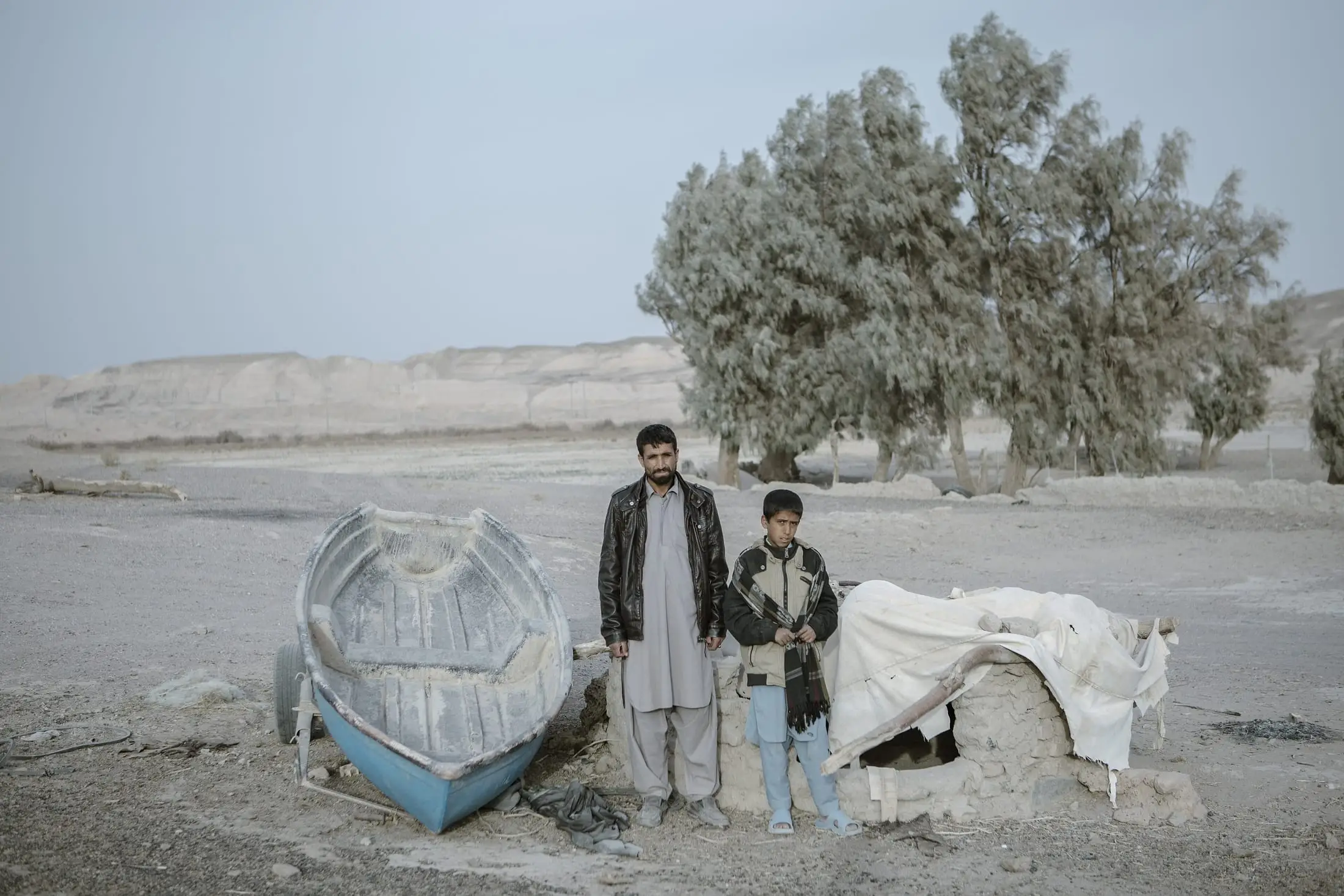مردی و پسری کنار قایق ماهیگیری‌ای ایستاده‌اند که روی زمین خشکِ جای تالاب هامون مانده است.